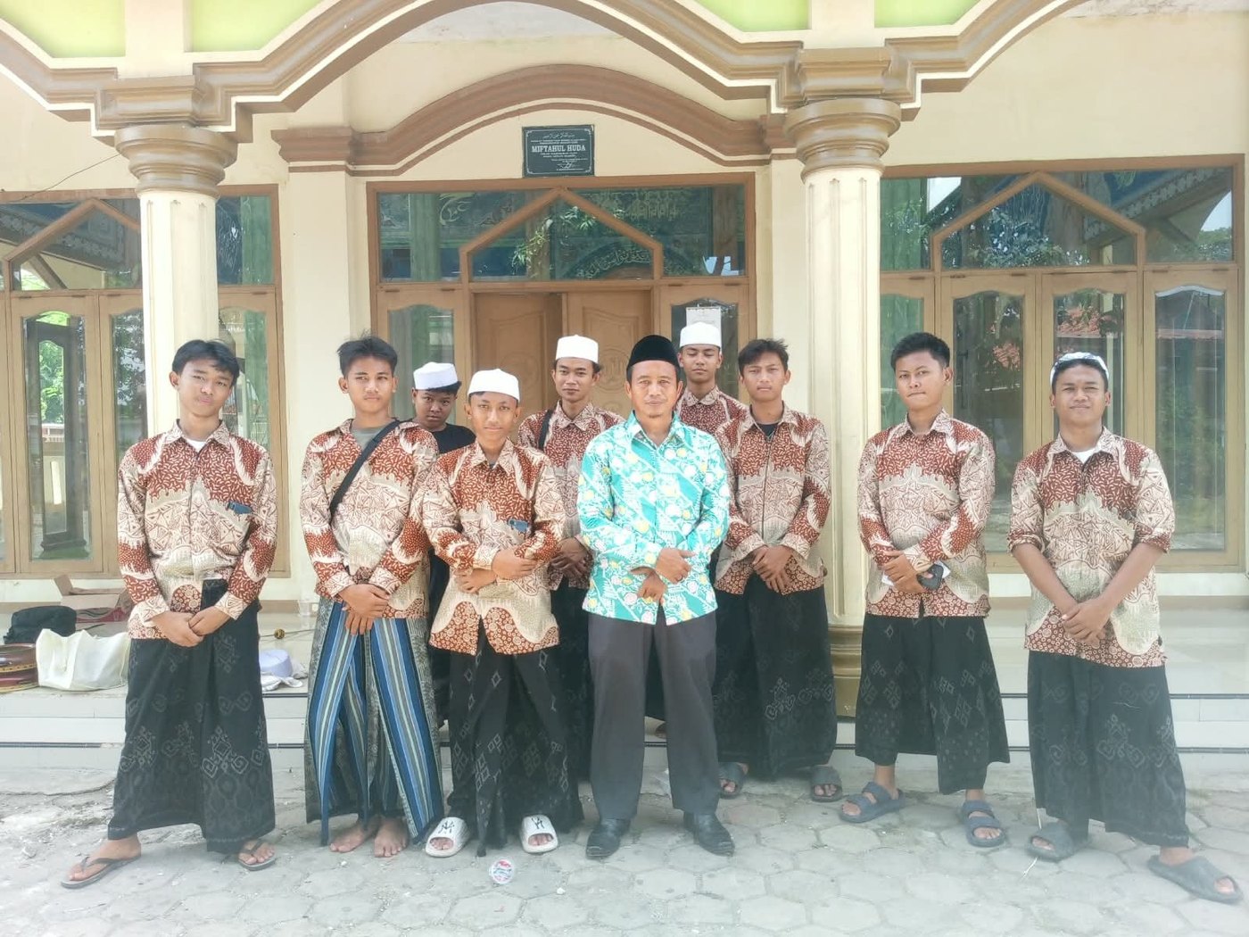 Foto grup hadroh Miftahul Huda Pakuning: para santri berkemeja batik dan berpeci putih berfoto bersama di depan bangunan pesantren, dengan rebana di sisi kiri